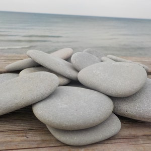 Puede incluir: Un conjunto de piedras de río grises y lisas de varios tamaños, sobre una superficie de madera desgastada. Las piedras son ovaladas y planas, con una forma natural y orgánica. El fondo muestra una masa de agua tranquila bajo un cielo nublado.