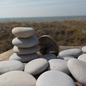 May include: A stack of smooth, gray stones balanced on top of each other, with a silver coin resting next to them. The stones are arranged on a wooden surface, with a blurred background of a sandy beach and ocean.