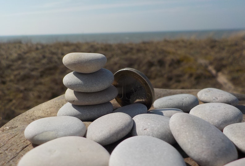 May include: A stack of smooth, gray stones balanced on top of each other, with a silver coin resting next to them. The stones are arranged on a wooden surface, with a blurred background of a sandy beach and ocean.