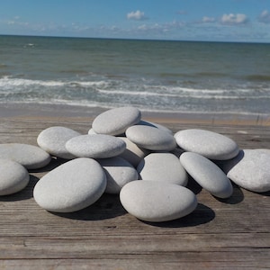 May include: A pile of smooth, white stones on a weathered wooden surface. The stones are various sizes and shapes, with a natural, polished finish. The background is a blurry image of a beach with blue water and sky.