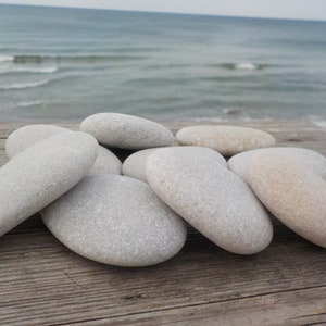 May include: A collection of smooth, gray beach stones of varying sizes. The stones are arranged on a weathered wooden surface, with the ocean and a sandy beach visible in the background. The stones have a natural, organic appearance.