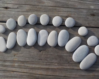 May include: A collection of smooth, grey stones arranged on a weathered wooden surface. The stones are in various shapes, from round to oblong, and are evenly spaced. The natural light enhances the texture of the wood and the stones.