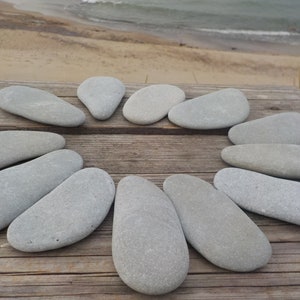 May include: A collection of smooth, gray river stones arranged in a circle on a weathered wooden surface. The stones vary in size and shape, with a natural, organic aesthetic. The background shows a beach and ocean.
