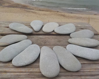 May include: A collection of smooth, gray river stones arranged in a circle on a weathered wooden surface. The stones vary in size and shape, with a natural, organic aesthetic. The background shows a beach and ocean.