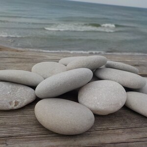May include: A pile of smooth, light gray stones on a weathered wooden surface. The stones are oval-shaped and have a natural, polished look.