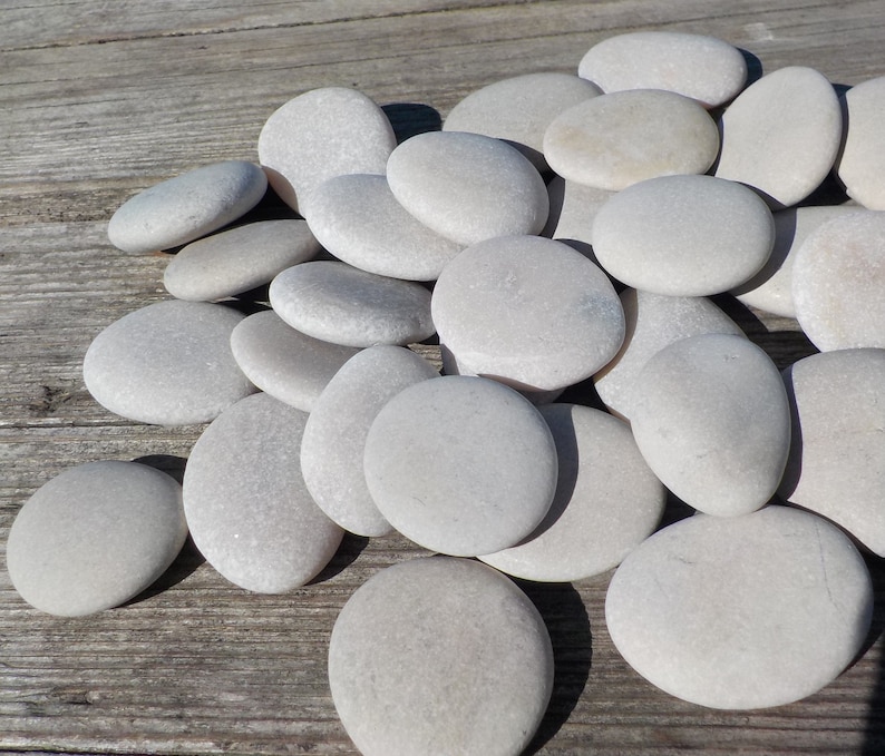 May include: A pile of smooth, light gray stones on a weathered wooden surface. The stones are various sizes and shapes, with a natural, polished finish.