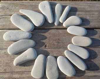 May include: A collection of smooth, gray river stones arranged in a circular pattern on a weathered wooden surface. The stones vary in shape, with some elongated and others more rounded. The natural light highlights the texture of the wood and the stones.