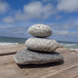 Può includere: Tre pietre lisce di colore grigio impilate l'una sull'altra su una superficie di legno. Le pietre sono rotonde e hanno una finitura naturale e testurizzata.