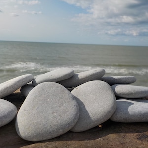 May include: A pile of smooth, light gray stones arranged on a wooden surface. The stones are various sizes and shapes, with a natural, weathered look. The background is a blurry image of a beach with waves crashing on the shore.