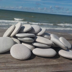 May include: A pile of smooth, white, oval-shaped stones on a wooden surface. The stones are arranged in a random pattern, with some overlapping others. The background is a blurry image of a beach with blue sky and white clouds.