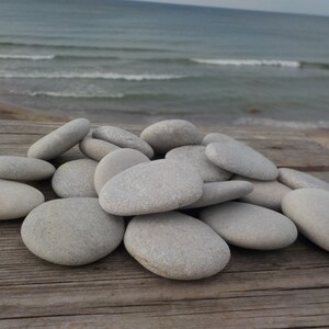 May include: A pile of smooth, light gray stones on a weathered wooden surface. The stones are various sizes and shapes, with some appearing more rounded than others.