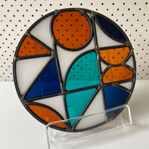 以下が含まれることがあります： 幾何学模様の丸いステンドグラス。オレンジ、青、ターコイズの形が特徴で、円や三角形など、黒い線で区切られています。ガラスは透明なスタンドに展示されています。