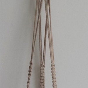 Puede incluir: Un macramé para plantas de color beige con una esfera de vidrio llena de pequeñas piedras y una vela blanca. El macramé tiene un lazo en la parte superior para colgarlo.
