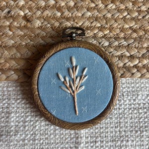 Puede incluir: Un pequeño bastidor de bordado redondo con tela azul claro y un diseño floral cosido. Tiene un marco de madera marrón y un cierre de metal. El fondo es una textura tejida, con un aspecto natural y rústico.