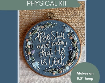 May include: A blue embroidery hoop with the text "Be Still and Know that He is God." The embroidery is surrounded by floral designs. The kit makes a 21.6 cm hoop. The text "Be Still and Know Embroidery PHYSICAL KIT" is at the top.