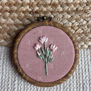 May include: A circular embroidery hoop frame with a faux wood grain finish containing dusty pink fabric. The fabric features a hand-stitched floral design with light pink petals and sage green stems, accented by small embroidered white cross stitches.