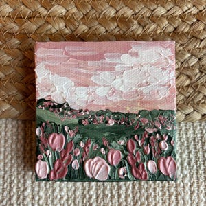 May include: Small square canvas art featuring a textured landscape painting. The artwork depicts a pink and white sky above a green field with pink flowers. The painting is set against a neutral woven background.