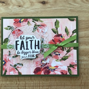 May include: Greeting card with a floral design in pink, red, and green. The card features the text "let your FAITH be bigger than your FEAR" in a white and black design, with a green ribbon tied around it.