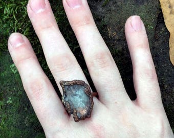 May include: A ring with a clear, faceted stone set in a copper setting. The stone is irregularly shaped and has a rough, natural look. The ring is worn on a person's finger.