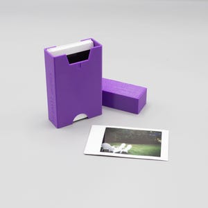 Può includere: Scatola di plastica viola per la conservazione di foto istantanee. La scatola ha una fessura per inserire le foto e un'etichetta con la scritta "INSTANT FILM STORAGE". Una foto è appoggiata accanto alla scatola.