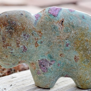 May include: A hand-carved stone sculpture of an elephant. The sculpture is primarily teal with brown and pink accents. The elephant has a rounded body and a small trunk. The stone has a rough, textured surface.