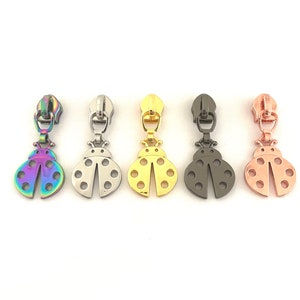 May include: Five different coloured metal zipper pulls shaped like ladybugs. The ladybugs are in shades of rainbow, silver, gold, black, and rose gold.