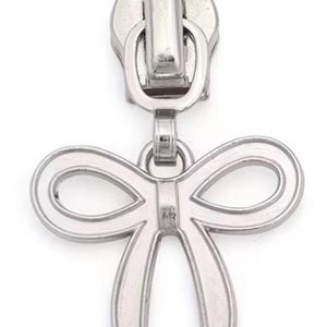 May include: A silver metal zipper pull featuring a bow-shaped charm. The zipper pull has a loop at the top for attachment. The bow charm is detailed with a double-loop design, suitable for various sewing projects.