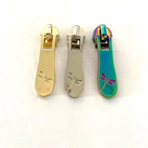 May include: Three zipper pulls featuring a dragonfly design. The pulls are gold, silver, and iridescent.
