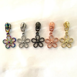 May include: Five zipper pulls with flower-shaped charms. The charms are in different colours: iridescent, silver, black, rose gold, and gold.
