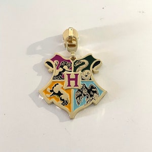May include: A gold-coloured zipper pull featuring the Hogwarts house crest. The crest is divided into four sections, each with a different animal and colour: a lion in gold, a snake in green, a badger in black, and an eagle in blue. The letter "H" is in the centre of the crest.