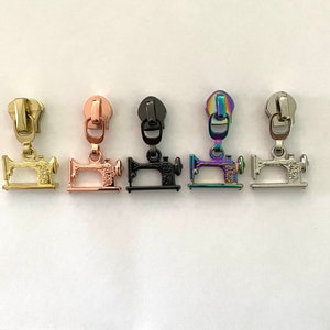May include: Five zipper pulls in different colours, each featuring a small sewing machine charm. The colours are gold, rose gold, black, iridescent, and silver.
