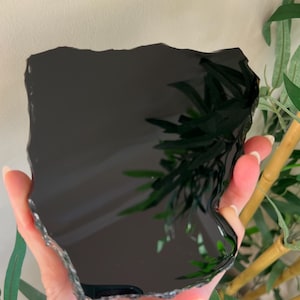 Op de afbeelding: Een gepolijste, platte, zwarte obsidiaan stenen plaat met onregelmatige, gekartelde randen. Het oppervlak is sterk reflecterend en heeft een heldere, spiegelachtige afwerking die licht en omringende vormen opvangt. De steen is donker en opaak.