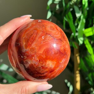 May include: A polished, spherical carnelian stone featuring a vibrant array of deep orange, burnt red, and translucent amber bands. The surface is smooth and glossy, showcasing intricate, swirling natural patterns throughout the entire orb.