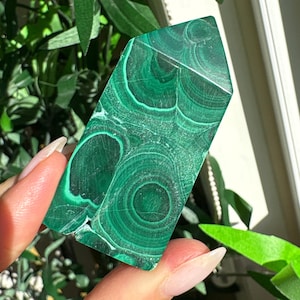 May include: A polished malachite crystal tower featuring a distinct banded pattern of deep forest green and lighter emerald green concentric circles. The stone is cut into a hexagonal prism shape with a pointed top, showcasing natural mineral layers.