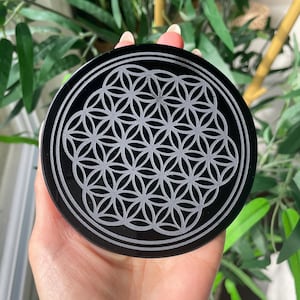 May include: Black obsidian coaster with a carved flower of life design. The coaster is round and has a polished surface.