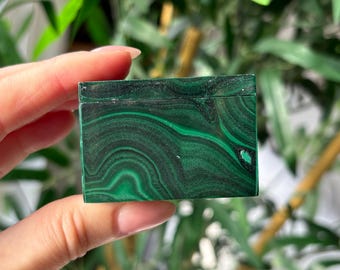 May include: A rectangular malachite box with a vibrant green color and swirling patterns. The polished surface reflects light, showcasing the natural beauty of the stone. The box is held in a hand, with a blurred background of green foliage.