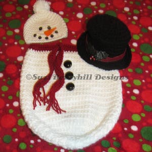 Op de afbeelding: Witte gehaakte baby outfit in de vorm van een sneeuwpop met een rode sjaal, een zwarte hoge hoed en zwarte knoopjesogen. De outfit is op een rode en groene stippenachtergrond.