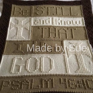 May include: A crocheted blanket featuring the words "Be Still and Know That I Am God" in brown and white yarn. The blanket has a brown border and a cross design in the centre.