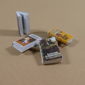 May include: Four miniature paperback books, including "Shadower" by Brenda Joyce, "The Renegade  Man" by Louis L'Amour, and "Santa  Fe" by  G.G.  Gregory. The books are on a brown surface with a gold coin.