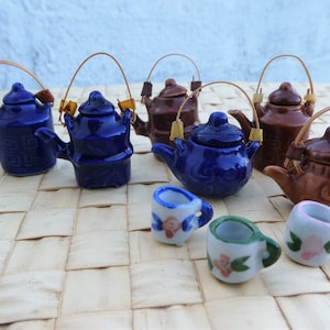 May include: A set of six miniature ceramic teapots in blue and brown with brown wooden handles. The teapots are arranged on a woven mat with three small ceramic teacups with floral designs.