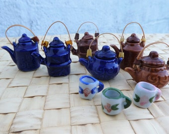 May include: A set of six miniature ceramic teapots in blue and brown with brown wooden handles. The teapots are arranged on a woven mat with three small ceramic teacups with floral designs.