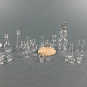 Puede incluir: Una colección de botellas de vidrio en miniatura, decantadores y vasos, que incluye una botella de vidrio con un dispensador de bomba, una cúpula de vidrio con una base de madera y una variedad de vasos para beber. Los artículos están dispuestos sobre una superficie gris.