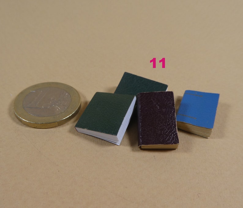 May include: Four miniature books, one green, one dark red, one blue, and one dark green, are arranged on a brown surface. The books are all about the same size and are smaller than a US one dollar coin.