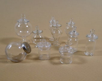 May include: A collection of clear glass jars with lids. The jars vary in shape and size, including round, cylindrical, and pedestal designs. Some jars have a small knob on top of the lid. The jars are arranged on a neutral background.