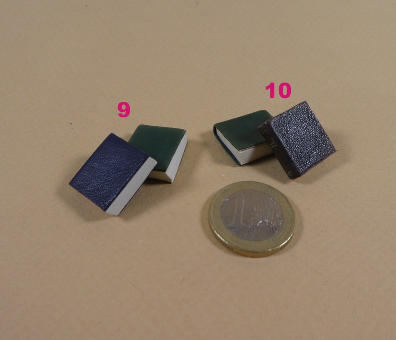 May include: Three miniature books, one blue, one green, and one brown, are arranged on a beige surface. A euro coin is in the background.