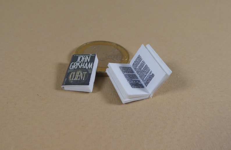 May include: A miniature paperback book titled "The Client" by John Grisham. The book is closed and lying on a brown surface. A coin is visible in the background.