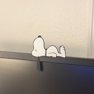 May include: A white and black Snoopy figure, in a relaxed pose, is perched on the edge of a black computer monitor. The cartoon character is lying down, with its head and feet visible. A black, teardrop-shaped piece hangs down.