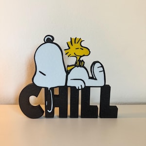 May include: A black and white Snoopy and Woodstock figure with the word "CHILL" in black block letters. Snoopy is lying down, and Woodstock is perched on his belly. The figure is made of wood and is set against a neutral background.