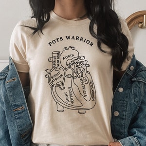 Puede incluir: Una camiseta beige con un contorno negro de un corazón humano con etiquetas anatómicas. El texto "POTS WARRIOR" está impreso en negro en la parte superior de la camiseta.