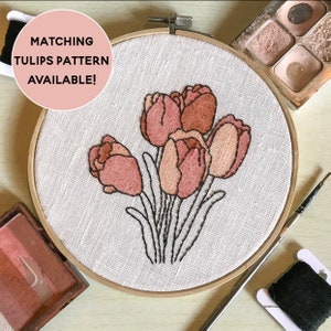 May include: A close-up of a hoop with a white fabric background featuring an embroidered design of a bouquet of pink and peach tulips. The hoop is surrounded by embroidery supplies, including a spool of black thread, a needle, and a small container of blush-colored makeup.